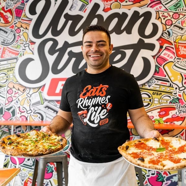 Smiling man in a black t-shirt holding two pizzas in front of a colorful Urban Street Pizza mural.