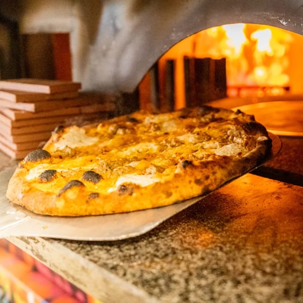 A pizza with a golden crust and melted cheese being pulled from a wood-fired oven.